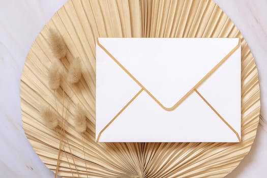 Blank envelope on dry beige palm leaf near hare's tail grass top view, mockup. Boho scene with envelope . Bohemian or close to nature greeting or wedding Invitation. Minimal composition.