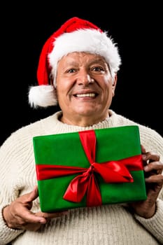 Shyly grinning old gentleman with a red Santa Claus cap and white knitted pullover. He is holding a green wrapped present with both hands in front of his chest. Gift giving theme. Isolated on black.