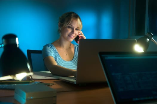 Beautiful woman working as interior designer, staying late at night in office with drawings and laptop computer to complete a project. The girl talks on mobile phone.