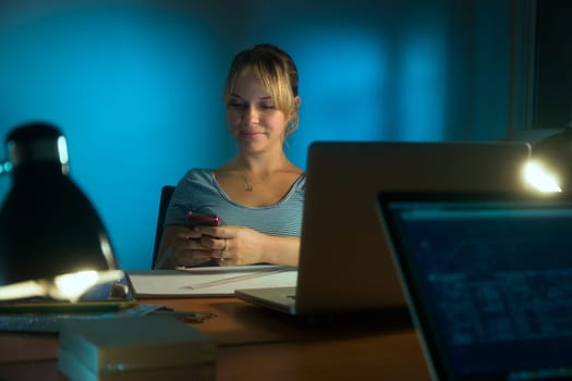 Beautiful woman working as interior designer, staying late at night in office with drawings and laptop computer to complete a project. The girl types a message on mobile phone.