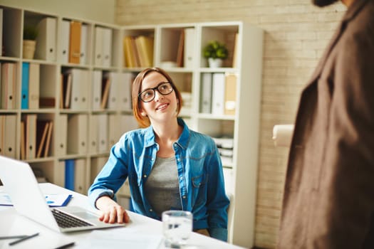 Pretty businesswoman sitting by workplace and listening to her colleague