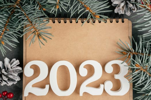 Happy New Year 2023. White numbers 2023 lying on folded notebook with Christmas tree branches and pine cones. Merry Christmas. Top view. Copy space