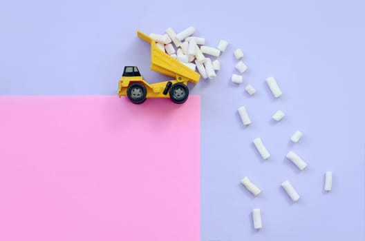 Yellow little toy dump truck throws marshmallow pieces from its raised back on a pastel violet and pink background. Flat lay minimal top view.