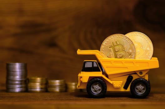 Yellow toy dumper loaded with shiny golden bitcoins is standing next to stacks of coins of various currencies on a wooden background. The advantage of digital currency versus physical money