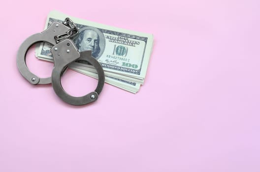 Police handcuffs and hundreds of us dollars lie on a background of living coral color. The concept of punishment for breaking the law in financial transactions
