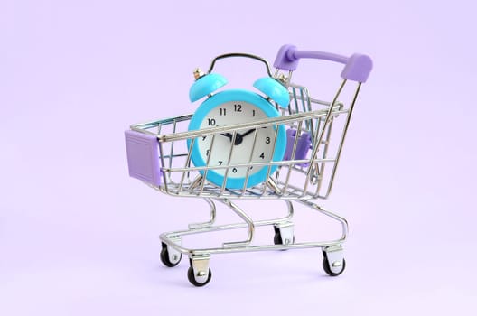 Blue alarm clock in supermarket trolley on violet background. Time resource acquisition concept. Time to sale