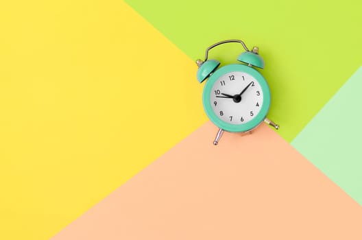 A small turquoise alarm clock is located between areas of yellow, peach and green colors. Time management concept. Flat lay minimal template with copy space. Top view