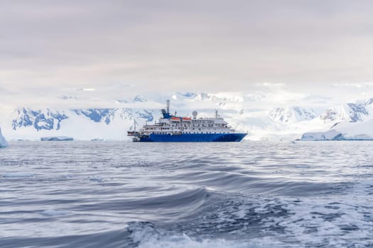 a blue, small expedition ship lies in front of an Antarctic iceberg landscape in Cierva Cove - a deep inlet on the west side of the Antarctic Peninsula, surrounded by rugged mountains and dramatic glacier fronts.