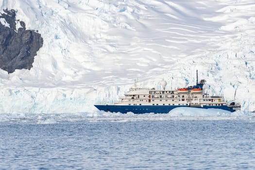 a blue, small expedition ship lies in front of an Antarctic iceberg landscape in Cierva Cove - a deep inlet on the west side of the Antarctic Peninsula, surrounded by rugged mountains and dramatic glacier fronts.