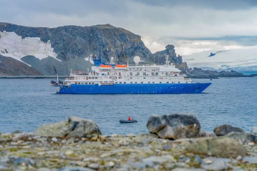 a blue, small expedition ship lies in front of an Antarctic iceberg landscape in Cierva Cove - a deep inlet on the west side of the Antarctic Peninsula, surrounded by rugged mountains and dramatic glacier fronts.