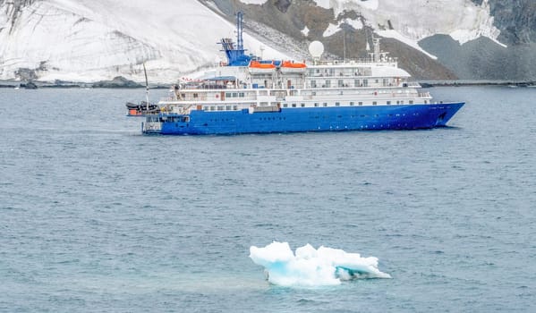 a blue, small expedition ship lies in front of an Antarctic iceberg landscape in Cierva Cove - a deep inlet on the west side of the Antarctic Peninsula, surrounded by rugged mountains and dramatic glacier fronts.