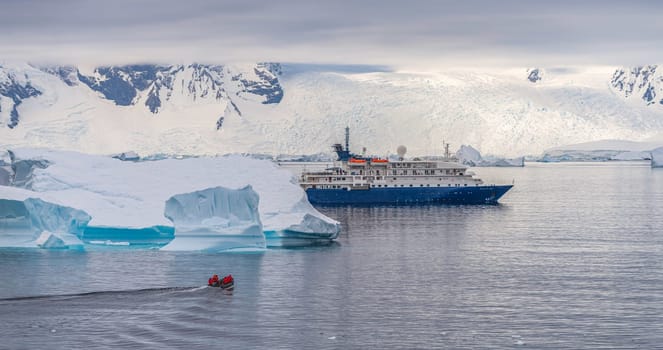 a blue, small expedition ship lies in front of an Antarctic iceberg landscape in Cierva Cove - a deep inlet on the west side of the Antarctic Peninsula, surrounded by rugged mountains and dramatic glacier fronts.