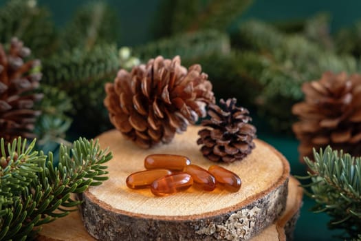 Orange softgel oil capsules near fir branches and pine cones on dark green close up. Preventive medicine and healthcare, taking dietary supplements and vitamins.