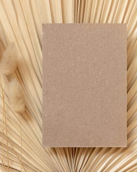 Blank card on dry beige palm leaf near hare's tail grass top view, mockup. Boho scene with kraft paper card. Bohemian or close to nature greeting or wedding Invitation. Minimal composition.