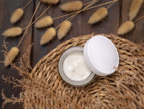 Opened cream jar with blank lid on wattled placemat on wood near Hare's Tail grass top view. Natural skincare product for everyday routine. Bohemian eco friendly cosmetics flat lay.
