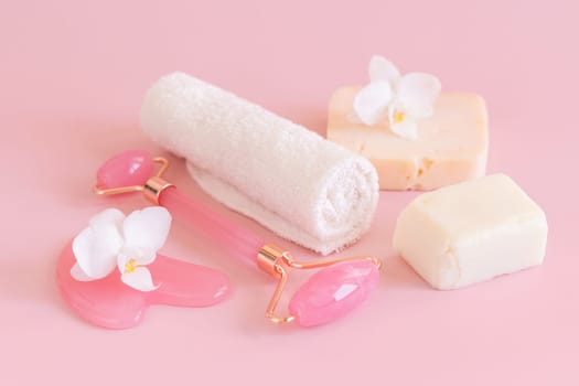 Facial roller, gua sha massager, towel and handmade soap bars near white orchid flower on pink close up. Anti-age massage and natural lifting therapy at home. Facial skin care at home. Beauty concept