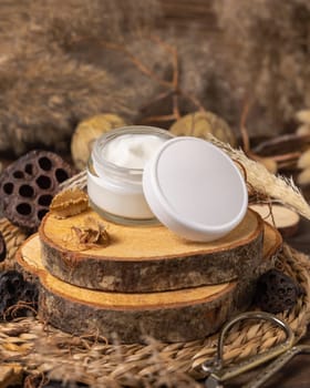 Opened glass cream jar with blank lid on wood near natural decorations close up, package mockup. Eco friendly skincare product, lotion or cream. Bohemian composition with dried leaves and flowers.