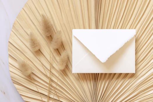 Square envelope on dry beige palm leaf near hare's tail grass top view, mockup. Boho scene with envelope . Bohemian or close to nature greeting or wedding Invitation. Minimal composition.