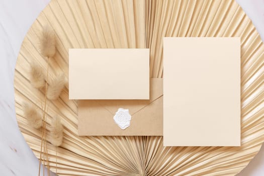 Blank cards and envelope on dry beige palm leaf near hare's tail grass top view, mockup. Boho scene with horizontal card. Bohemian or close to nature greeting card or wedding Invitation.