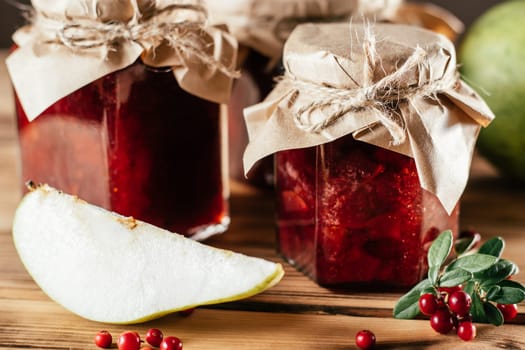 Jars of homemade lingonberry and pear jam with craft paper on lids on wooden surface next to fresh lingonberries and pears.