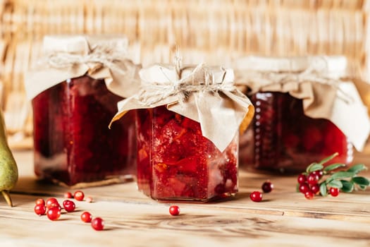 Jars of homemade lingonberry and pear jam with craft paper on lids on wooden surface next to fresh lingonberries and pears.