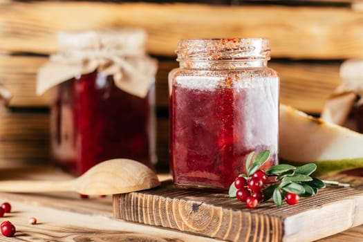 Jars of homemade lingonberry and pear jam with craft paper on lids on wooden surface next to fresh lingonberries and pears.