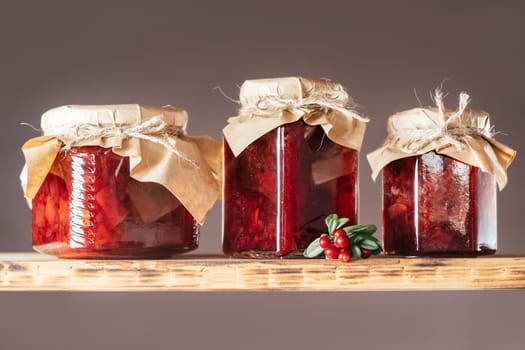 Jars of homemade lingonberry and pear jam with craft paper on lids on wooden shelf next to fresh lingonberries on brown background