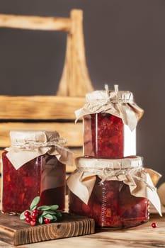 Jars of homemade lingonberry and pear jam with craft paper on lids on wooden surface, next to fresh lingonberries. Vertical