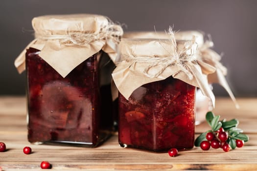 Jars of homemade lingonberry and pear jam with craft paper on lids on wooden surface next to fresh lingonberries