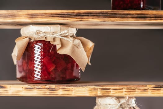 Jar of homemade lingonberry and pear jam with craft paper on lid on wooden shelf on brown background