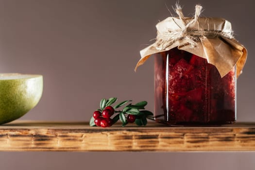 Jar of homemade lingonberry and pear jam with craft paper on lid on wooden shelf next to fresh lingonberries and pears on brown background.