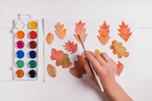Female hand drawing autumn leaves of different shapes, sizes and colors with watercolors and brush on white wooden table. Top view. DIY handmade autumn decorations