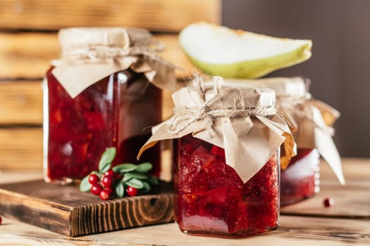 Jars of homemade lingonberry and pear jam with craft paper on lids on wooden surface next to fresh lingonberries and pears.