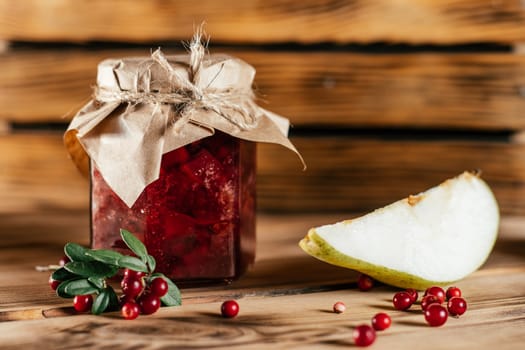 Jar of homemade lingonberry and pear jam with craft paper on lid on wooden surface next to fresh lingonberries and pears.