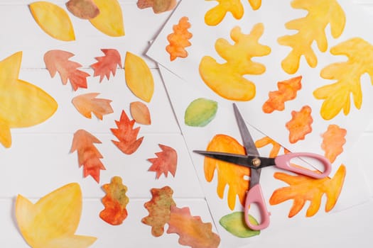 Drawn on white paper autumn leaves of different shapes, sizes and colors with scissors. Cut out leaves on white wooden table. top view. DIY handmade autumn decorations
