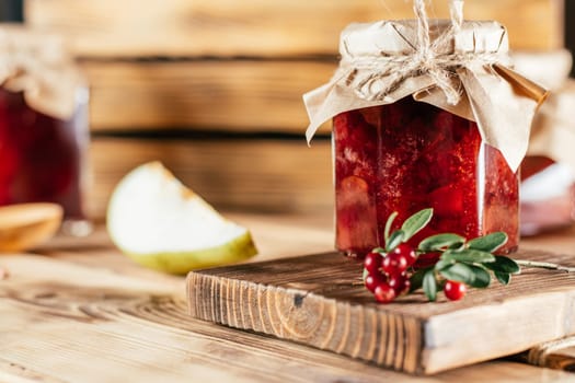 Jar of homemade lingonberry and pear jam with craft paper on lid on wooden surface next to fresh lingonberries and pears.