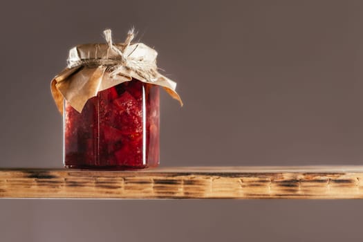 Jar of homemade lingonberry and pear jam with craft paper on lid on wooden shelf on brown background
