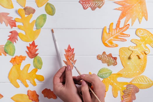 Female hand drawing autumn leaves of different shapes, sizes and colors with watercolors and brush on white wooden table. Top view. DIY handmade autumn decorations