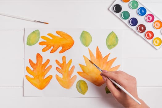Female hand drawing autumn leaves of different shapes, sizes and colors with watercolors and brush on white wooden table. Top view. DIY handmade autumn decorations