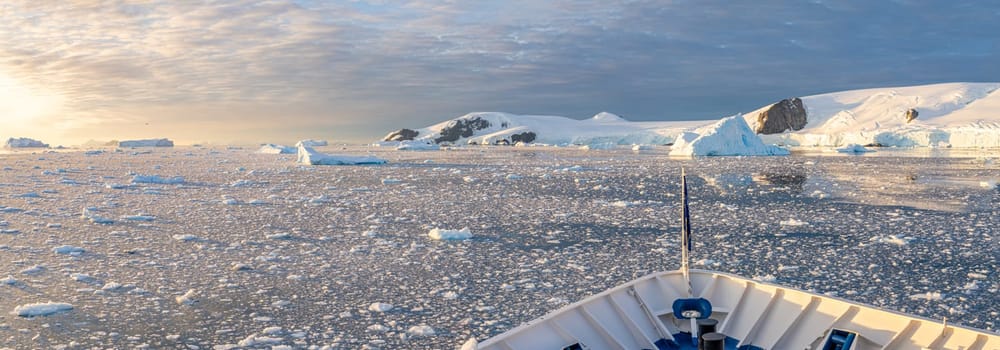 Cruise ship sails through wintry Cierva Cove - a deep inlet on the west side of the Antarctic Peninsula, ringed by Cierva Bay in San Martín Land - Antarctica, during a dramatic sunset / evening sunset.
