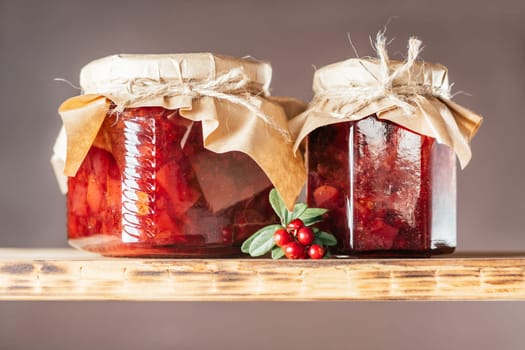 Jars of homemade lingonberry and pear jam with craft paper on lids on wooden shelf next to fresh lingonberries on brown background