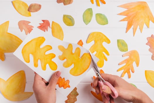 Female hands cutting out with scissors drawn on white paper autumn leaves of different shapes, sizes and colors. Cut out leaves on white wooden table. top view. DIY handmade autumn decorations