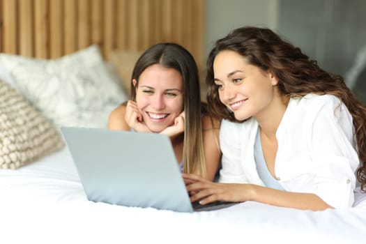Two happy women watching media on laptop lying on a bed in the bedroom