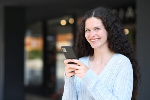 Happy woman holds smart phone and looks at camera standing in the street