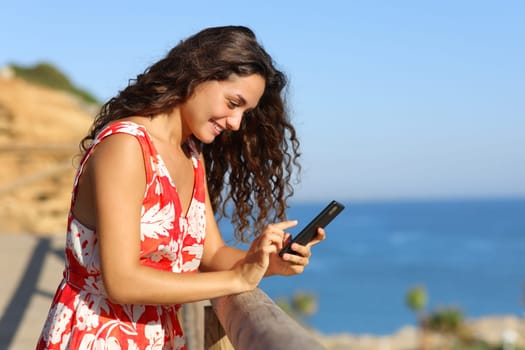 Side view portrait of a happy woman on summer vacation on the beach using phone