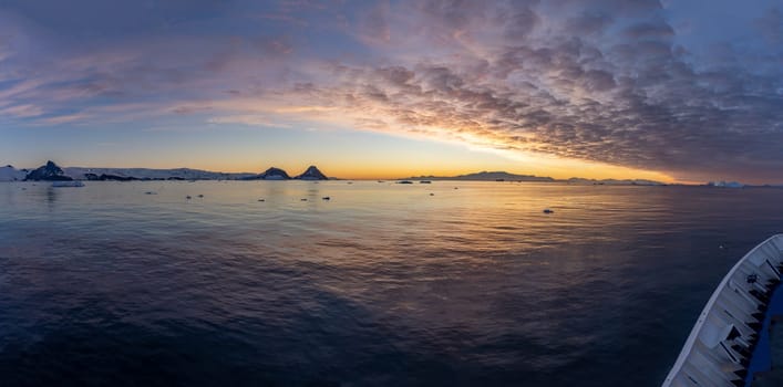 Cruise ship sails through wintry Cierva Cove - a deep inlet on the west side of the Antarctic Peninsula, ringed by Cierva Bay in San Martín Land - Antarctica, during a dramatic sunset / evening sunset.