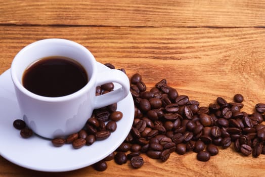 Freshly made coffee in a white Cup on a white saucer surrounded by scattered coffee beans on a wooden table. Fresh roasted coffee. Place for an inscription or logo.