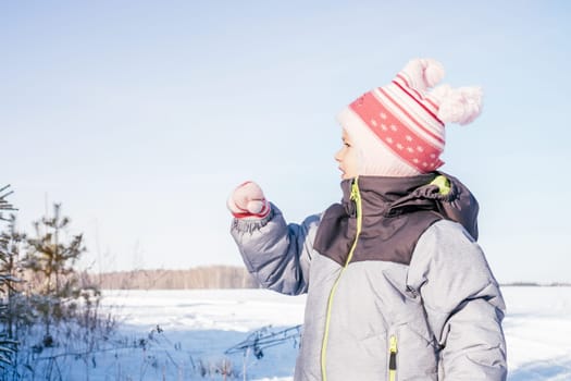 Little girl 3-4 years old in winter clothes overalls, hat and mittens, stands outdoors against clear blue sky and fir trees and looks away, pointing with hand. Winter holidays, children outdoors