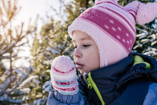 Little girl 3-4 years old in winter clothes overalls, hat and mittens, stands outdoors against snow-covered pines and fir trees and eats snow on sunny day. Winter holidays, children playing outdoors