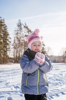 Little girl 3-4 years old in winter overalls, hat and mittens, stands outdoors against against blue sky and fir trees and throws snow over her laughing. Winter holidays, children outdoors. Vertical